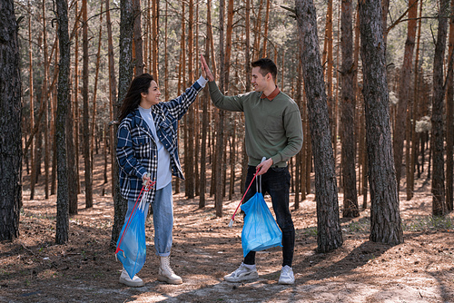 full length of happy couple with trash bags giving high five while picking up rubbish with grabber tools in forest