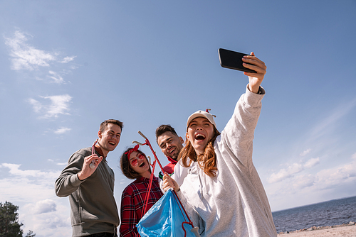 cheerful woman taking selfie with friends while picking up trash
