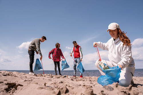happy woman holding trash bag and picking up rubbish on sand near group of volunteers
