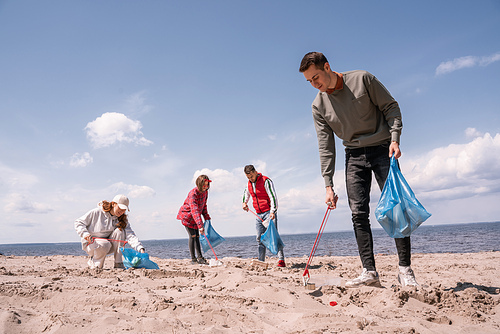 happy man holding trash bag and picking up rubbish on sand near group of volunteers