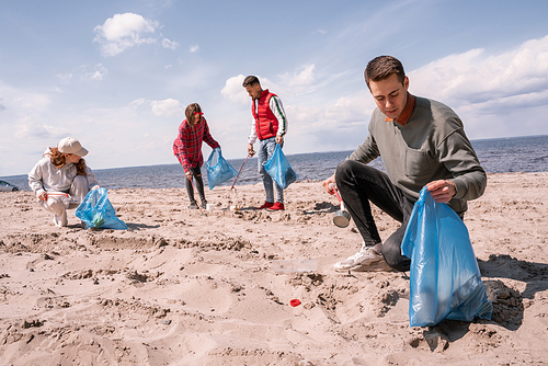 young man holding trash bag and collecting rubbish on sand near group of volunteers