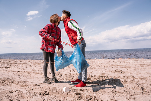 couple with trash bags and grabbers kissing while standing on sand