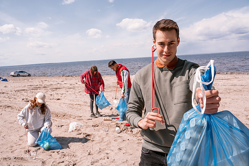 happy man holding trash bag near group of blurred volunteers picking up rubbish on sand