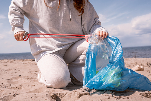 cropped view of woman holding trash bag and picking up rubbish on sand