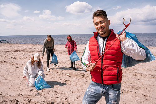 cheerful man holding trash bag and grabber near blurred volunteers collecting rubbish on sand