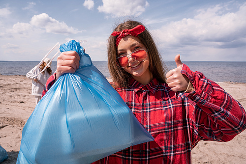happy young woman in sunglasses holding trash bag and showing thumb up, ecology concept