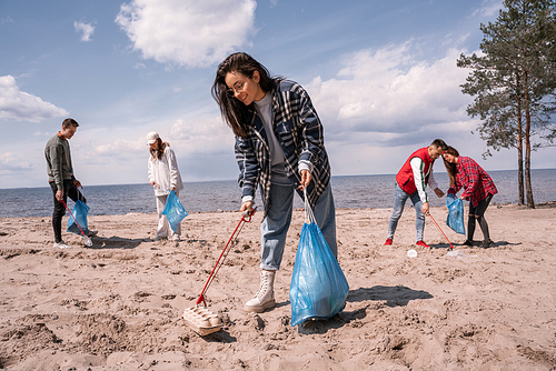 smiling young woman holding trash bag and collecting rubbish on sand near group of volunteers