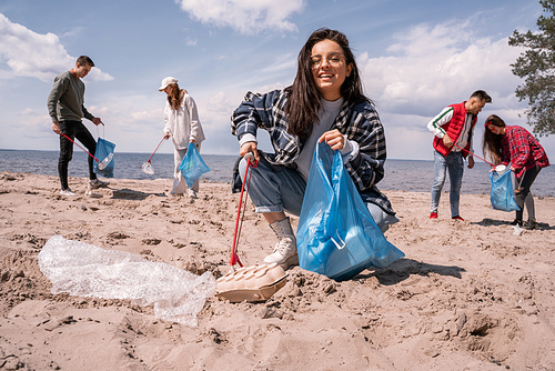 happy young woman collecting rubbish with grabber near group of volunteers
