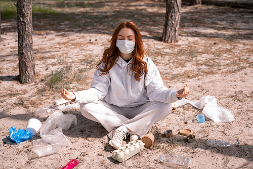 young woman in medical mask meditating near trash on ground