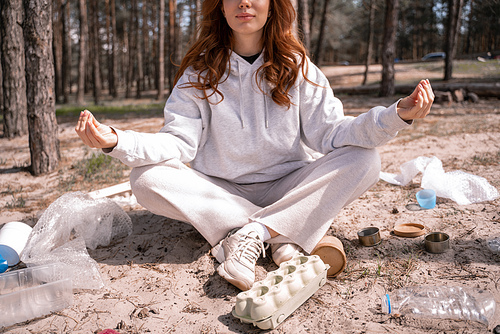 cropped view of young woman with crossed legs sitting and meditating near trash on ground