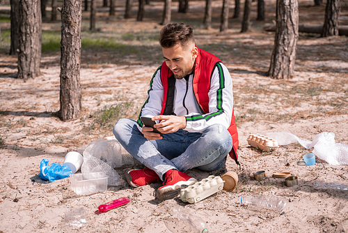 cheerful man with crossed legs sitting and using smartphone near trash on ground