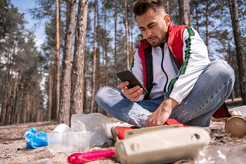 man with crossed legs sitting and using smartphone near trash on ground