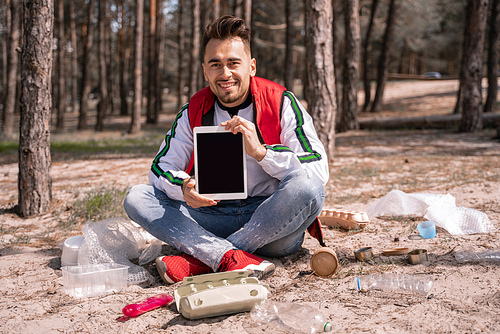 happy man with crossed legs sitting and holding digital tablet with blank screen near trash on ground