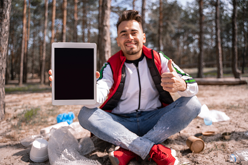 happy man with crossed legs holding digital tablet with blank screen and showing thumb up near trash on ground