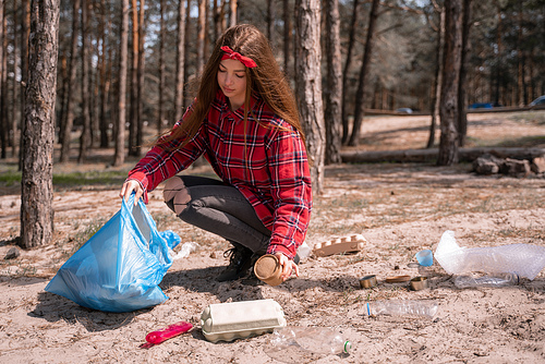 young woman holding trash bag and collecting rubbish on ground in forest