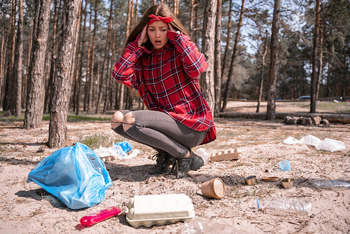 shocked young woman looking at garbage near trash bag in forest