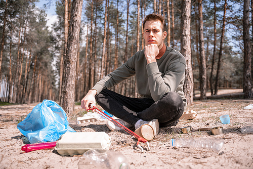 pensive man with crossed legs sitting near garbage on ground in woods