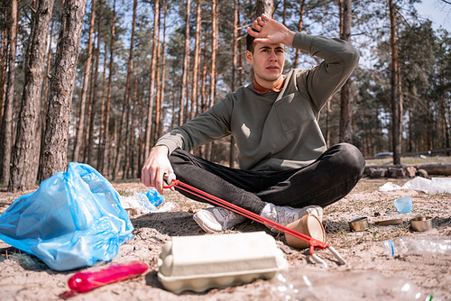 tired volunteer sitting near trash bag and rubbish in forest