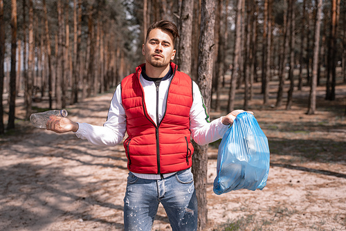 young man holding blue trash bag and plastic bottle in forest