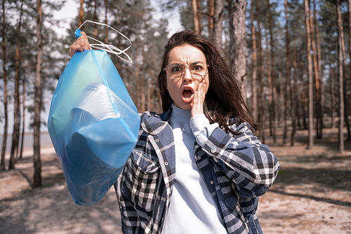 shocked young woman holding blue trash bag in forest