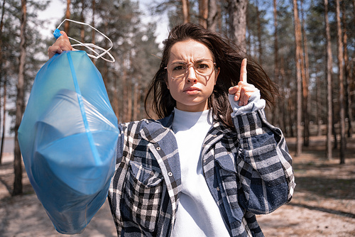 young woman warning while pointing with finger and holding blue trash bag in forest
