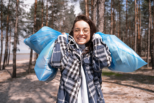 cheerful young woman in glasses holding blue trash bags in forest