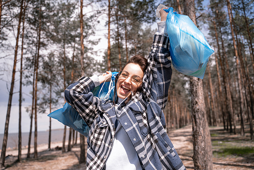 excited young woman in glasses holding blue trash bags in forest
