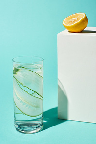 transparent glass with pure water and cucumber slices near lemon half on turquoise background