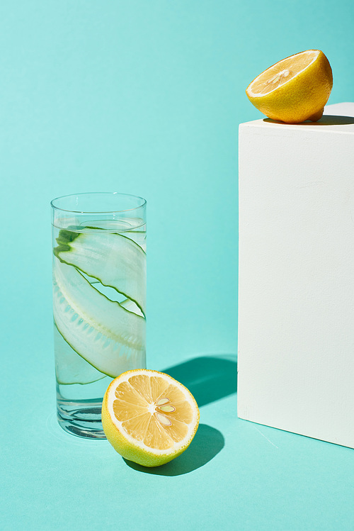 transparent glass with pure water and cucumber slices near lemons on turquoise background