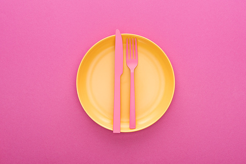 top view of pink plastic fork and knife on yellow plastic plate on pink background