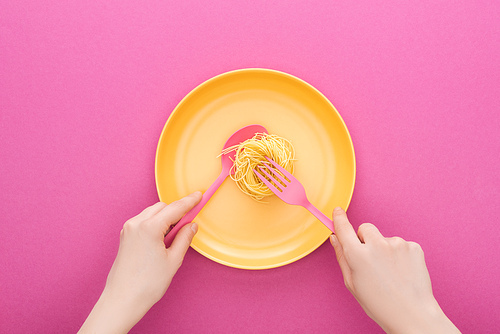 cropped view of adult holding pink spoon and taking vermicelli pasta with fork on yellow plastic plate on pink background