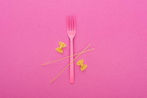 uncooked farfalle pasta near pink plastic fork and spaghetti isolated on pink