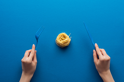 cropped view of woman holding blue plastic fork and knife with vermicelli pasta in middle on blue background