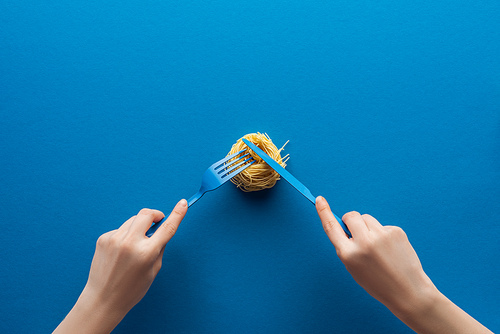 cropped view of woman holding blue plastic fork and cutting vermicelli pasta with knife on blue background