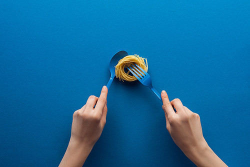 cropped view of woman holding blue spoon and taking vermicelli pasta with fork isolated on blue