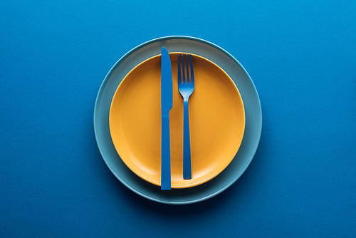 top view of blue plastic knife and fork on yellow plate above another plate on blue background