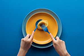 cropped view of woman holding blue spoon and taking vermicelli pasta with fork on yellow plate above another plate on blue background