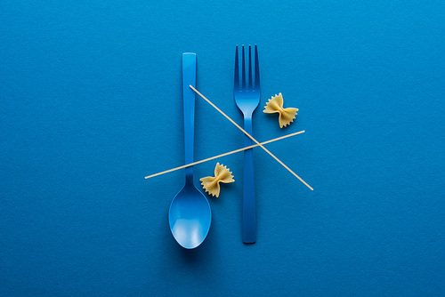 top view of spaghetti on blue plastic spoon upside down and fork near farfalle pasta on blue background