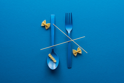 top view of spaghetti on blue plastic spoon upside down and fork near two kinds of pasta on blue background