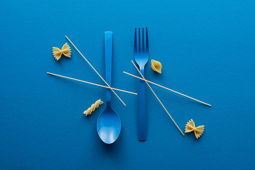 flat lay of spaghetti on blue plastic spoon upside down and fork near different kinds of uncooked pasta on blue background