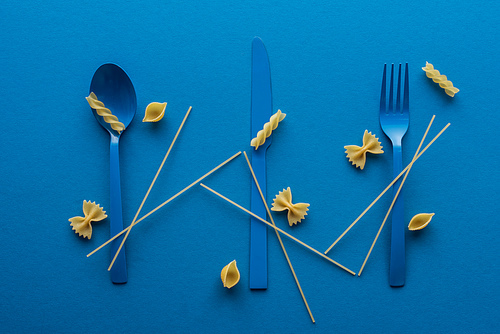 blue plastic cutlery with uncooked spaghetti and different kinds of pasta on blue background