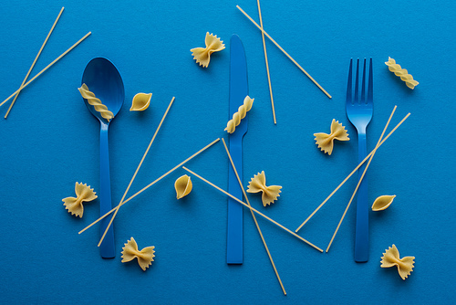 blue plastic cutlery with uncooked spaghetti around and different kinds of pasta on blue background