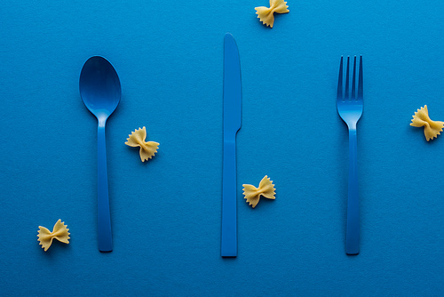 blue plastic spoon, knife and fork with uncooked farfalle pasta around on blue background