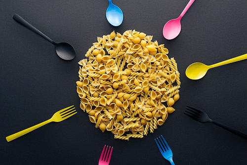 top view of uncooked pasta surrounded by colorful cutlery on black background
