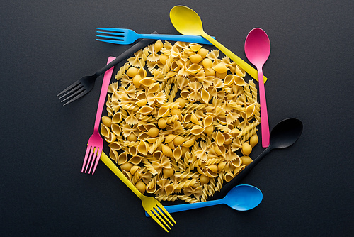 top view of uncooked pasta surrounded by colorful spoons and forks on black background