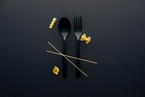 black plastic spoon, fork and four different kinds of pasta on black background