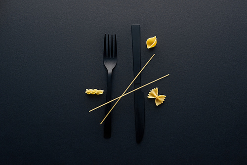 black plastic knife upside down, fork and four different kinds of pasta on black background