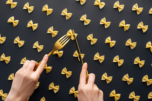 cropped view of woman holding yellow fork and cutting farfalle pasta with knife isolated on black