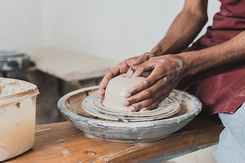 partial view of young african american man modeling wet clay on wheel with hands in pottery