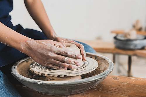 partial view of young african american woman sitting on bench and modeling wet clay on wheel with hands in pottery